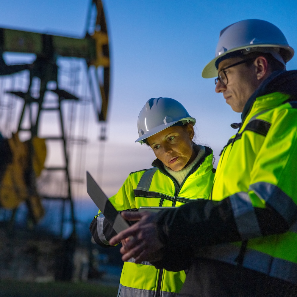 Two workers in yellow hazard jackets at an oil field looking at laptop